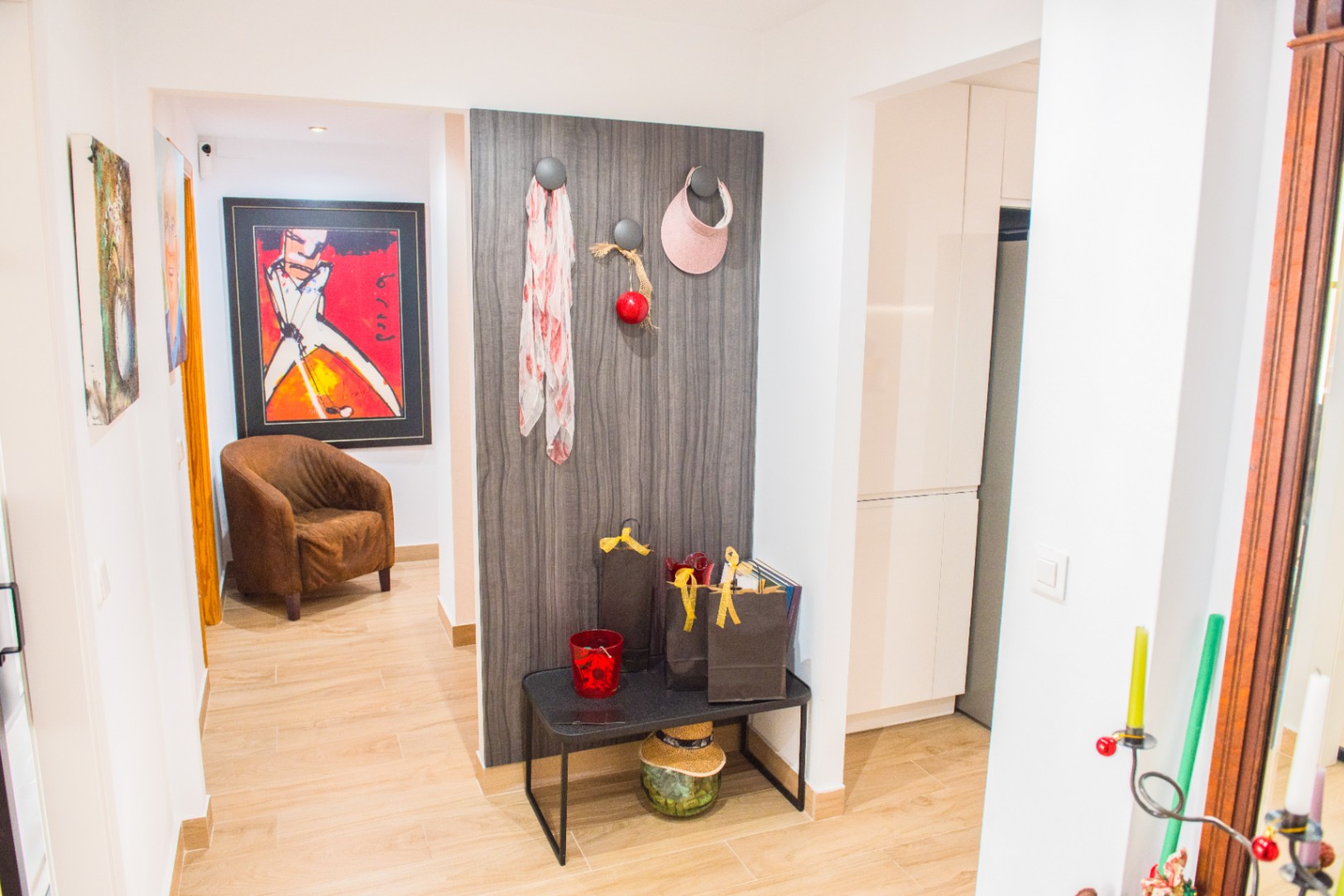 Entrée avec sol en grès cérame effet bois, panneau mural décoratif sombre et banc minimaliste noir. Le couloir attenant présente des œuvres d'art et un fauteuil brun.