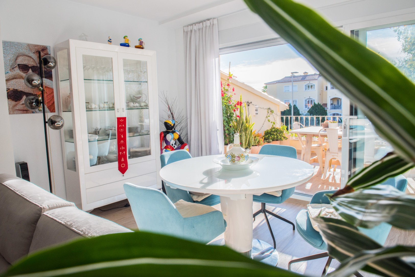 Salle à manger lumineuse avec table ronde blanche, chaises en velours bleu et vitrine d'exposition, s'ouvrant sur une terrasse ensoleillée.