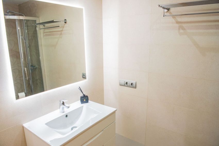 Salle de bain moderne avec meuble-lavabo blanc, miroir éclairé et murs carrelés de couleur beige. Une cabine de douche vitrée est visible en reflet. Salle de bain moderne avec meuble-lavabo blanc, miroir éclairé et murs carrelés de couleur beige. Une cabine de douche vitrée est visible en reflet.