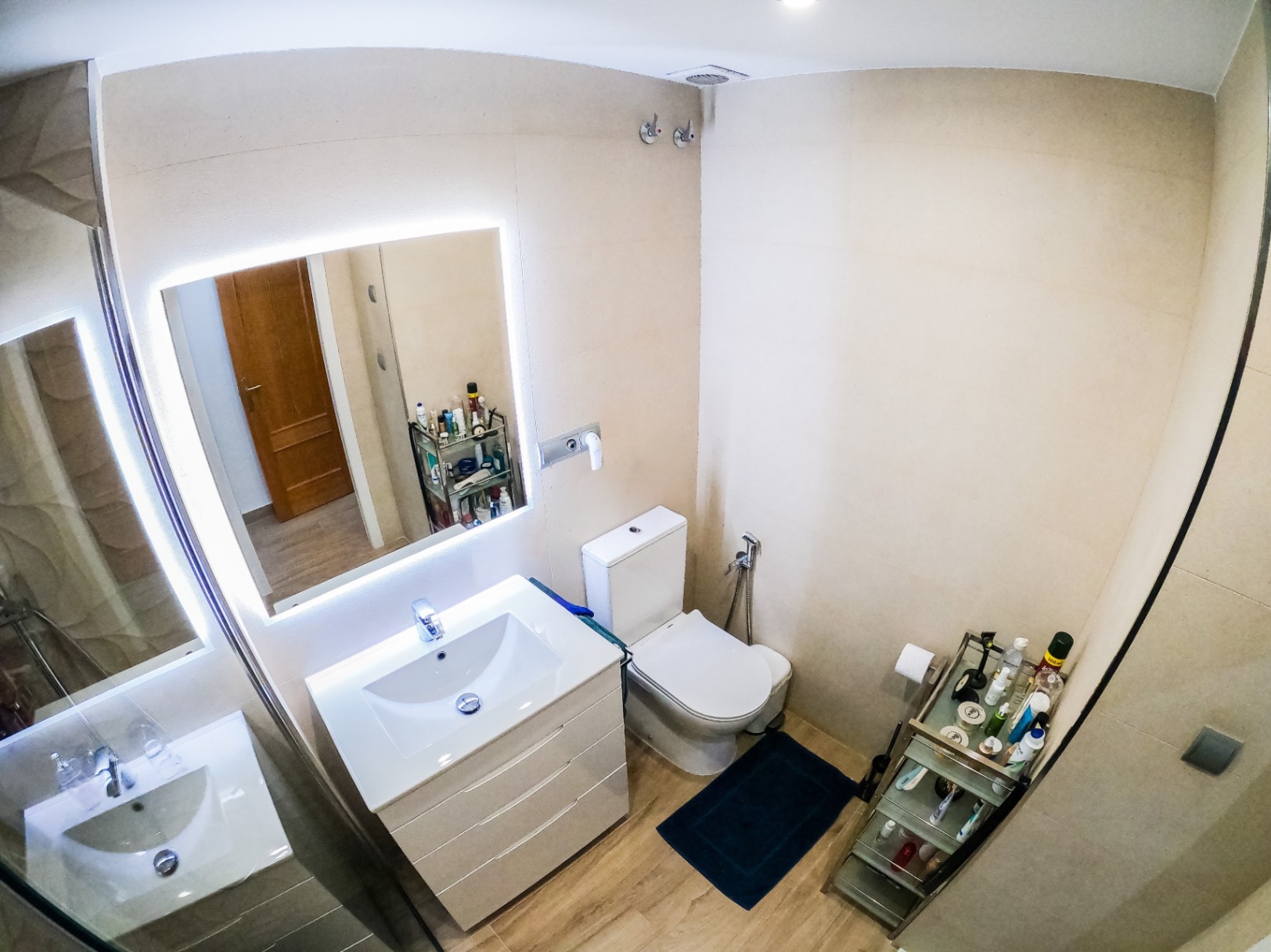 Salle de bain moderne avec meuble vasque blanc, miroir rétroéclairé par LED et sol imitation bois. Murs carrelés en beige et toilettes avec douchette intégrée.