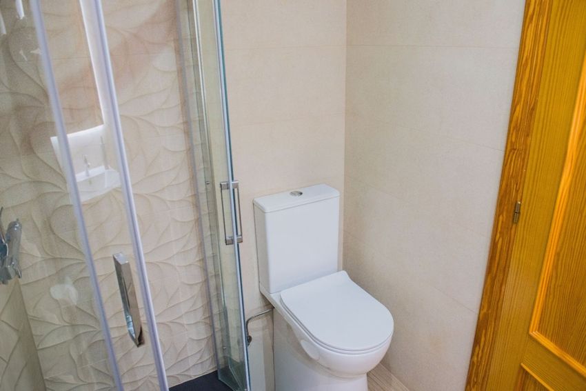 Salle de bain moderne présentant des toilettes blanches et une cabine de douche vitrée avec poignées chromées. Le carrelage mural beige est texturé de motifs de feuilles dans la douche. Salle de bain moderne présentant des toilettes blanches et une cabine de douche vitrée avec poignées chromées. Le carrelage mural beige est texturé de motifs de feuilles dans la douche.