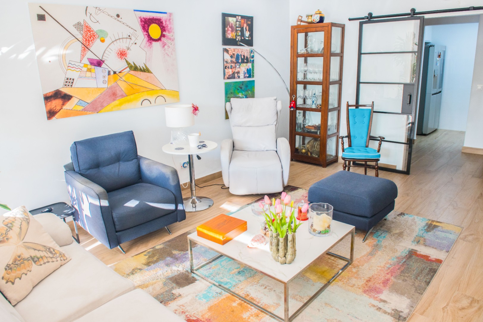 Salon avec fauteuil bleu, relax blanc et vitrine en bois. Décoré d'un tableau abstrait, d'un tapis coloré et d'une porte coulissante vitrée au cadre noir industriel.