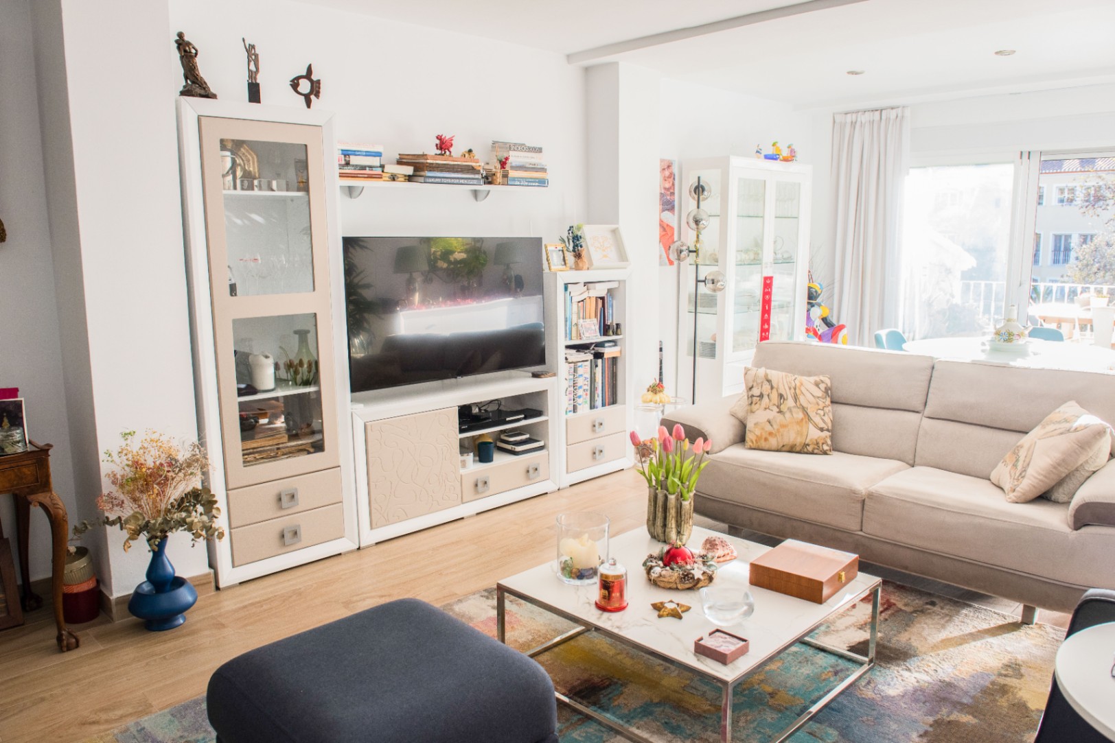 Salon lumineux avec canapé en tissu beige, table basse en marbre et meuble TV modulaire blanc. De grandes baies vitrées offrent une lumière naturelle abondante.
