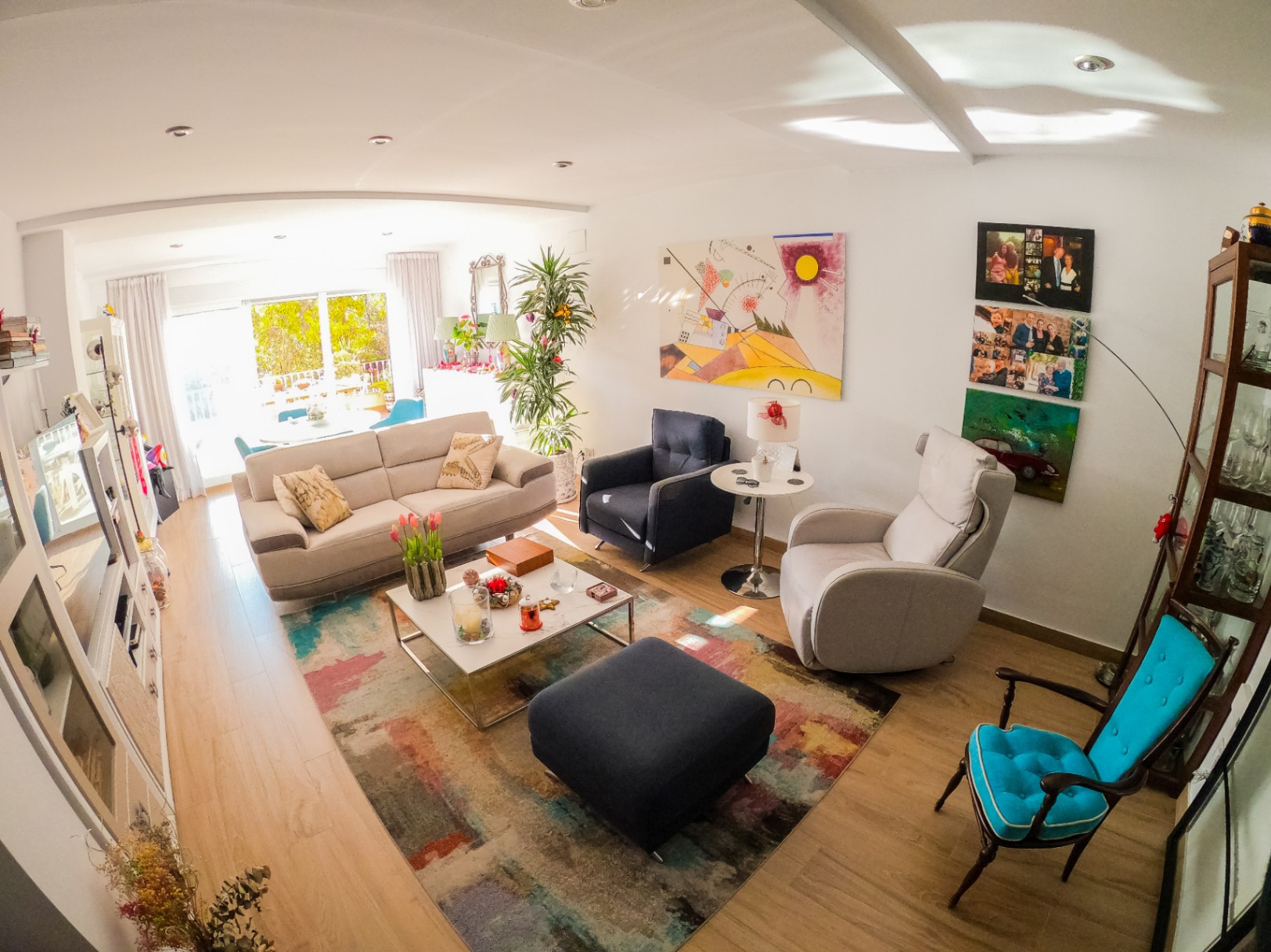 Salon lumineux avec parquet clair, tapis abstrait multicolore et mobilier varié. De grandes baies vitrées offrent une lumière naturelle et un accès à une terrasse verdoyante.