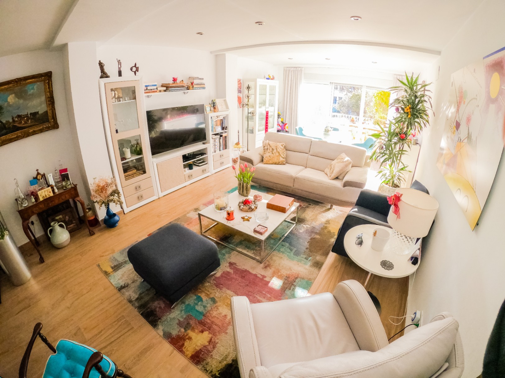 Salon spacieux avec parquet clair, tapis abstrait coloré et canapé beige. De grandes baies vitrées coulissantes apportent une luminosité naturelle et ouvrent sur un balcon.