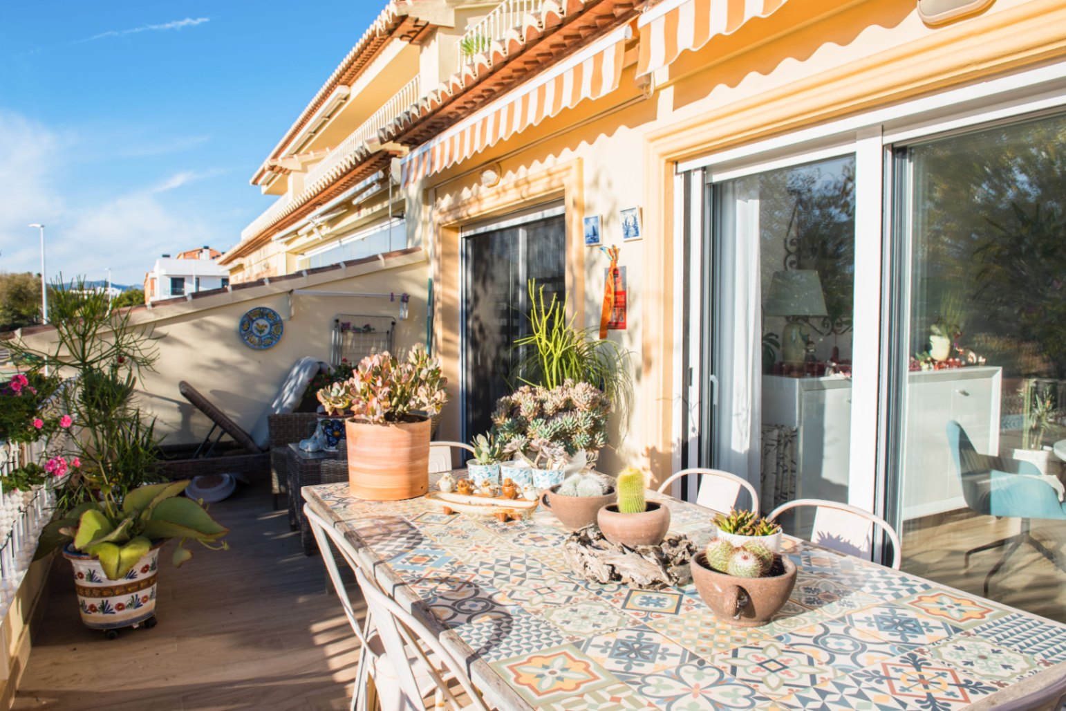 Terrasse méditerranéenne ensoleillée avec table à manger en mosaïque, plantes succulentes et grandes baies vitrées sous un store banne rayé.