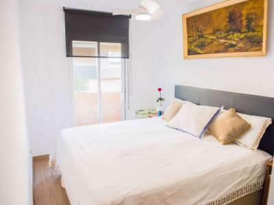 Chambre avec lit double, tête de lit grise capitonnée, parquet en bois et grande fenêtre avec store foncé. Ventilateur de plafond et tableau paysager complètent l'espace.