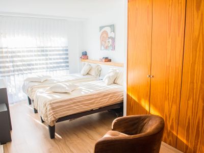 Chambre lumineuse avec deux lits simples, une armoire en bois naturel et un sol clair. La lumière naturelle pénètre par les stores verticaux, éclairant la pièce.