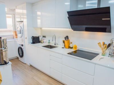 Cuisine moderne avec armoires blanches laquées, plaque à induction noire et hotte inclinée. L'espace intègre un éclairage sous les meubles et des appareils de buanderie près de la fenêtre.