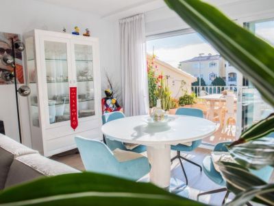 Espace repas lumineux avec table ronde blanche moderne et chaises bleu clair. De grandes portes vitrées coulissantes mènent à une terrasse végétalisée, offrant lumière naturelle et vues résidentielles.