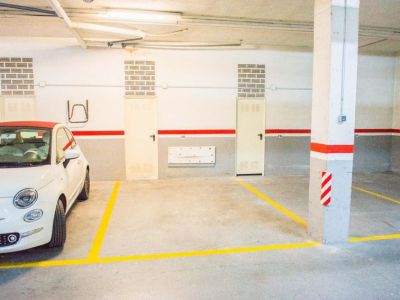 Place de parking souterrain spacieuse avec marquages jaunes clairs sur sol en béton. Comprend un éclairage aérien, des piliers et des unités de rangement.