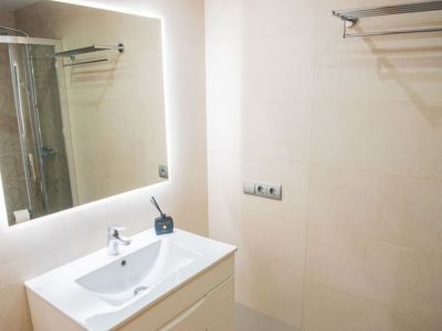 Salle de bain moderne avec meuble-lavabo blanc, miroir éclairé et murs carrelés de couleur beige. Une cabine de douche vitrée est visible en reflet.