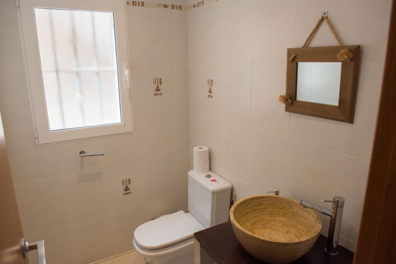 Cabinet de toilette avec vasque en pierre naturelle sur meuble sombre, miroir suspendu par une corde rustique et murs carrelés clairs avec fenêtre dépolie.