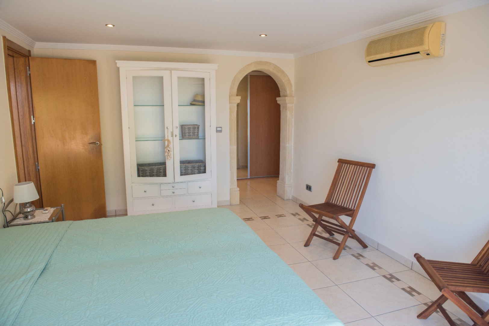 Chambre avec arche en pierre, sol carrelé avec bordure décorative, vitrine blanche et chaises pliantes en bois. Équipée d'une climatisation murale et de spots encastrés.