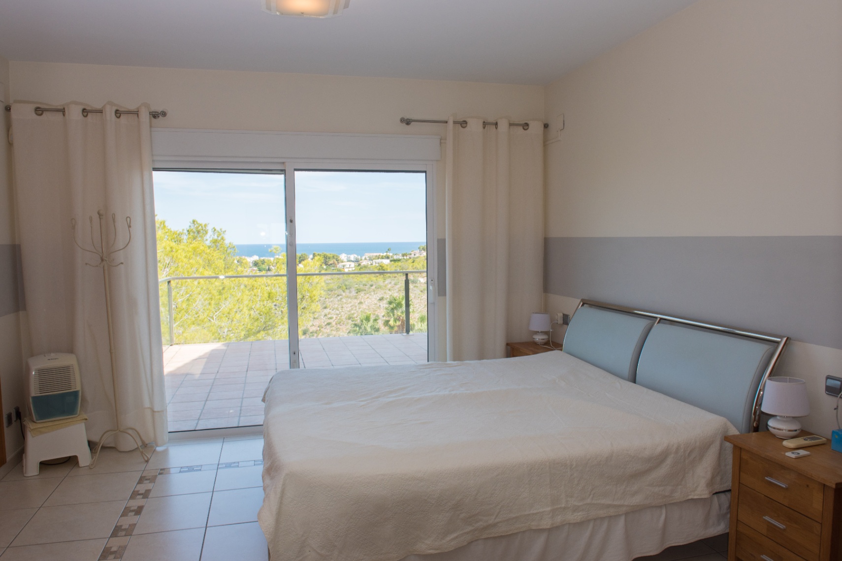 Chambre avec carrelage au sol et baies vitrées donnant sur une terrasse avec vue mer. Comprend une tête de lit métallique moderne et des murs bicolores.