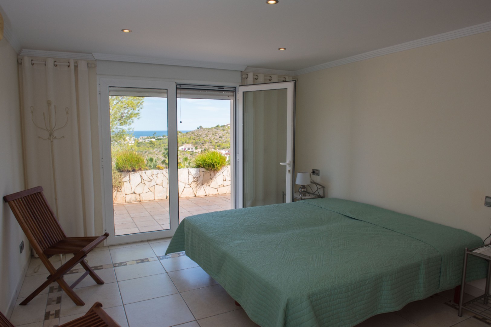 Chambre lumineuse avec accès direct à une terrasse carrelée offrant une vue sur la mer et les collines, complétée par un éclairage à spots encastrés.
