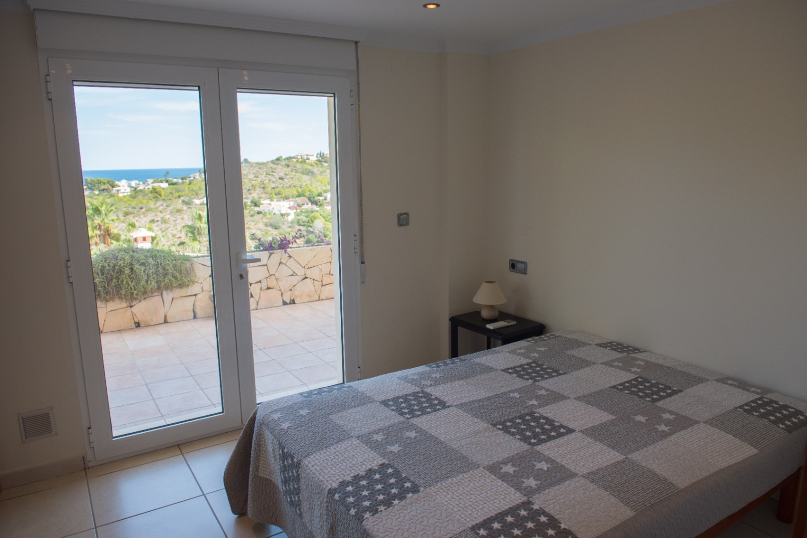Chambre lumineuse avec accès direct à une terrasse carrelée par des portes vitrées, offrant une vue dégagée sur la mer et les collines environnantes.