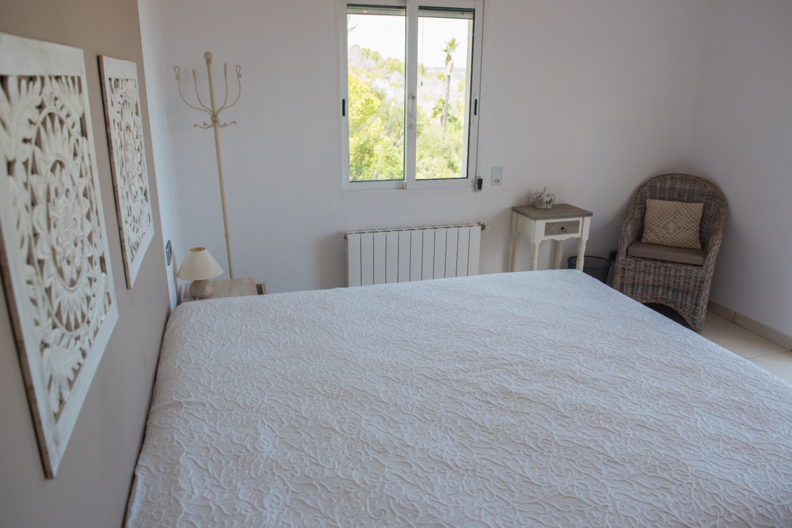 Chambre lumineuse avec couvre-lit blanc texturé, décorations murales sculptées et fauteuil en osier. La fenêtre donne sur une végétation méditerranéenne.