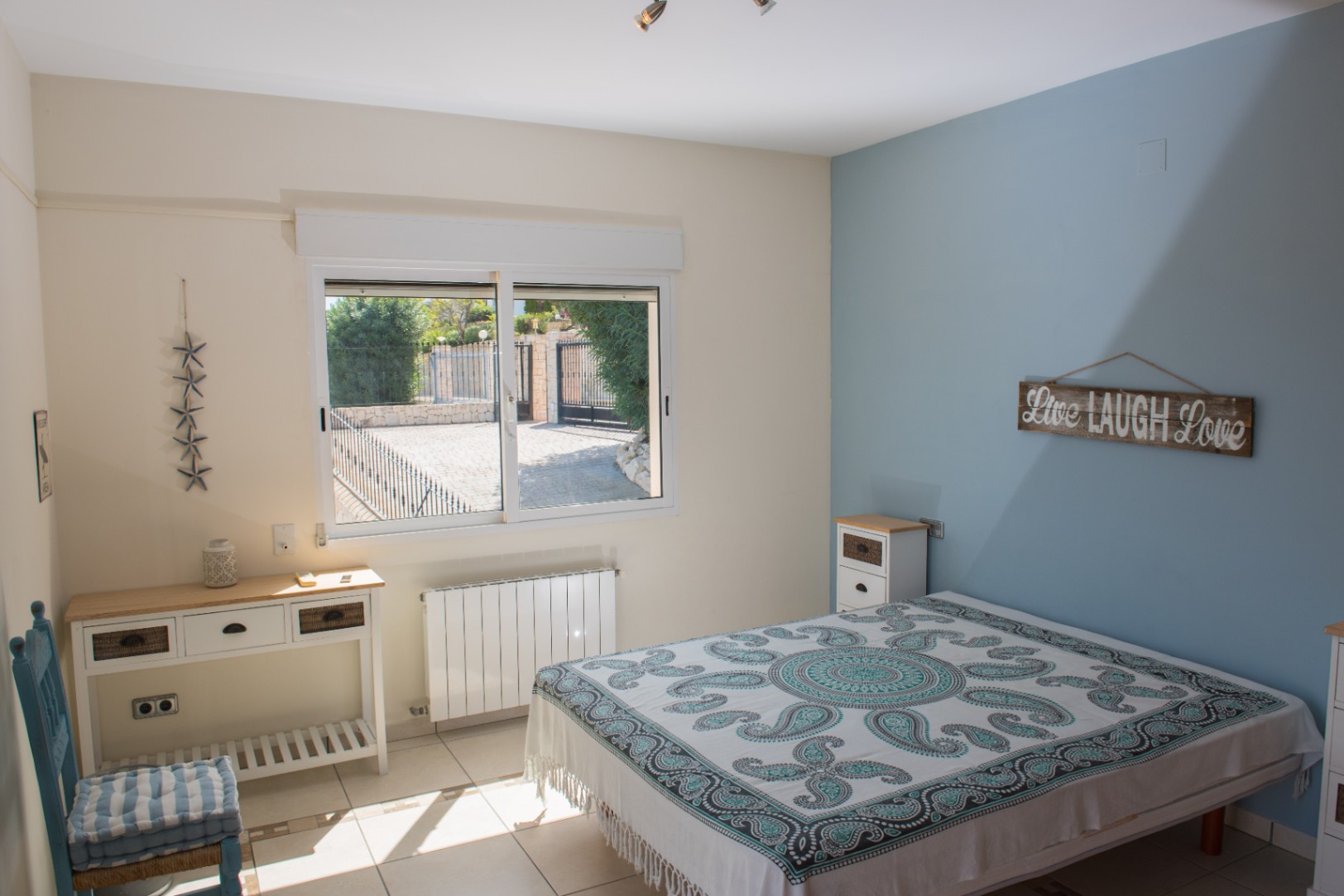 Chambre lumineuse avec mur d'accent bleu, sol carrelé et fenêtre offrant une vue sur l'allée pavée sécurisée. Comprend une console blanche et un radiateur.