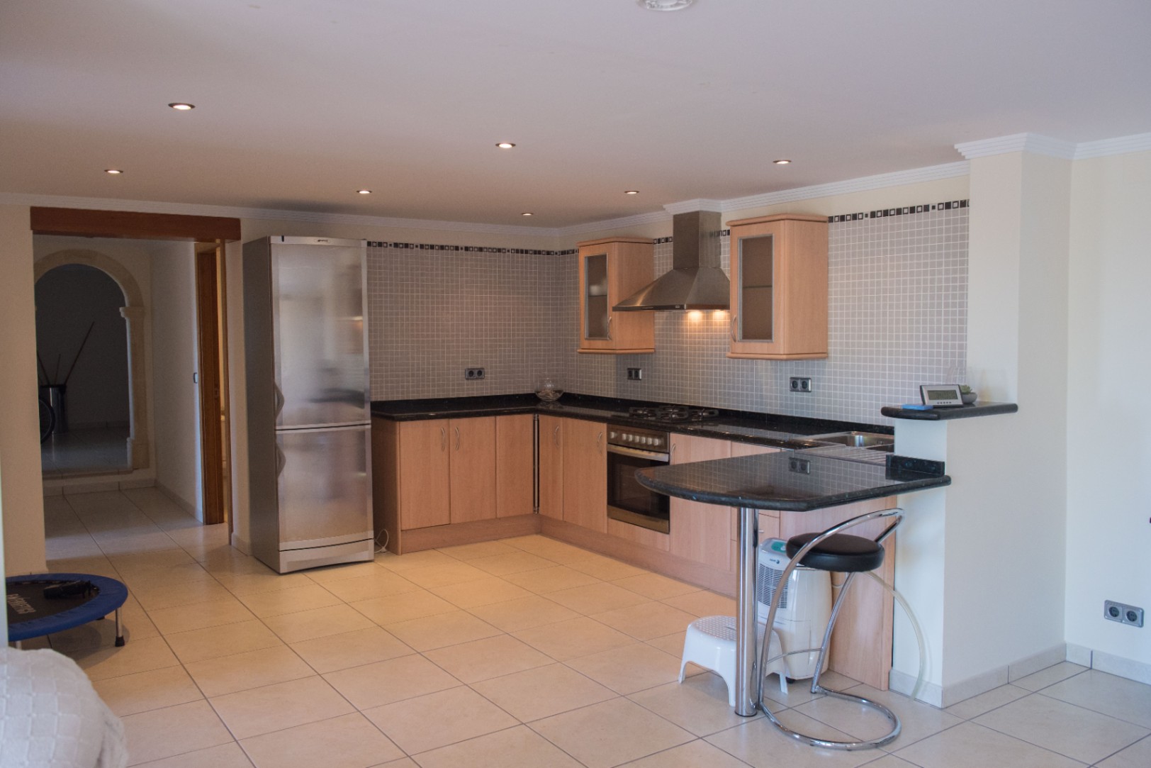 Cuisine en L dotée de meubles en bois clair, plans de travail en granit noir et électroménager en inox. Comprend un coin repas, une crédence en mosaïque et des spots intégrés.