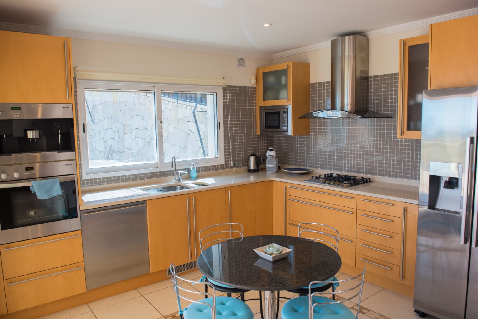 Cuisine moderne avec placards en bois clair, appareils Miele en inox et crédence en mosaïque grise. Comprend une table en granit noir et un plan de travail blanc épuré.