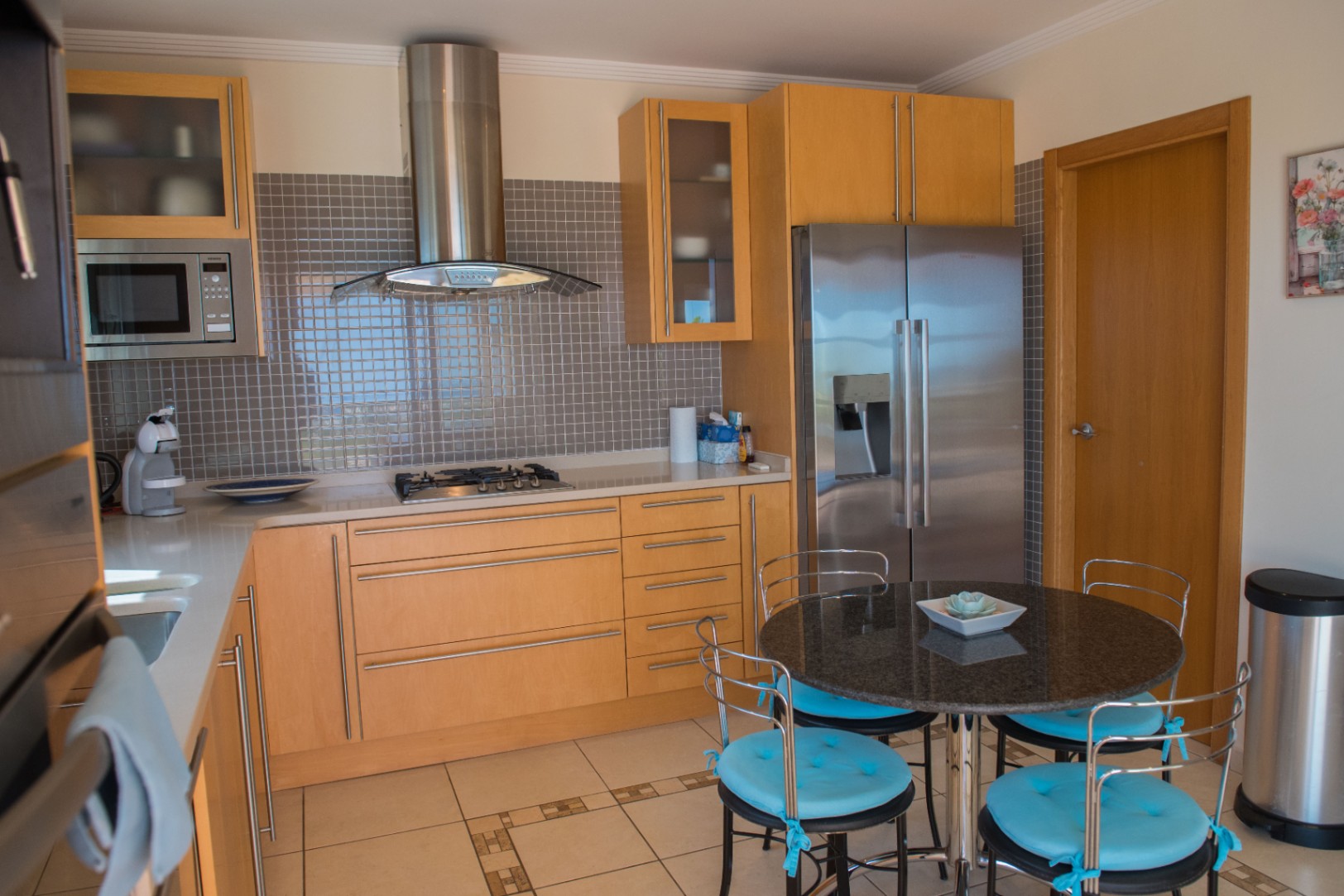 Cuisine moderne avec placards en bois clair, crédence en mosaïque grise et électroménager en inox. Comprend une table ronde en granit et des chaises aux coussins bleus.