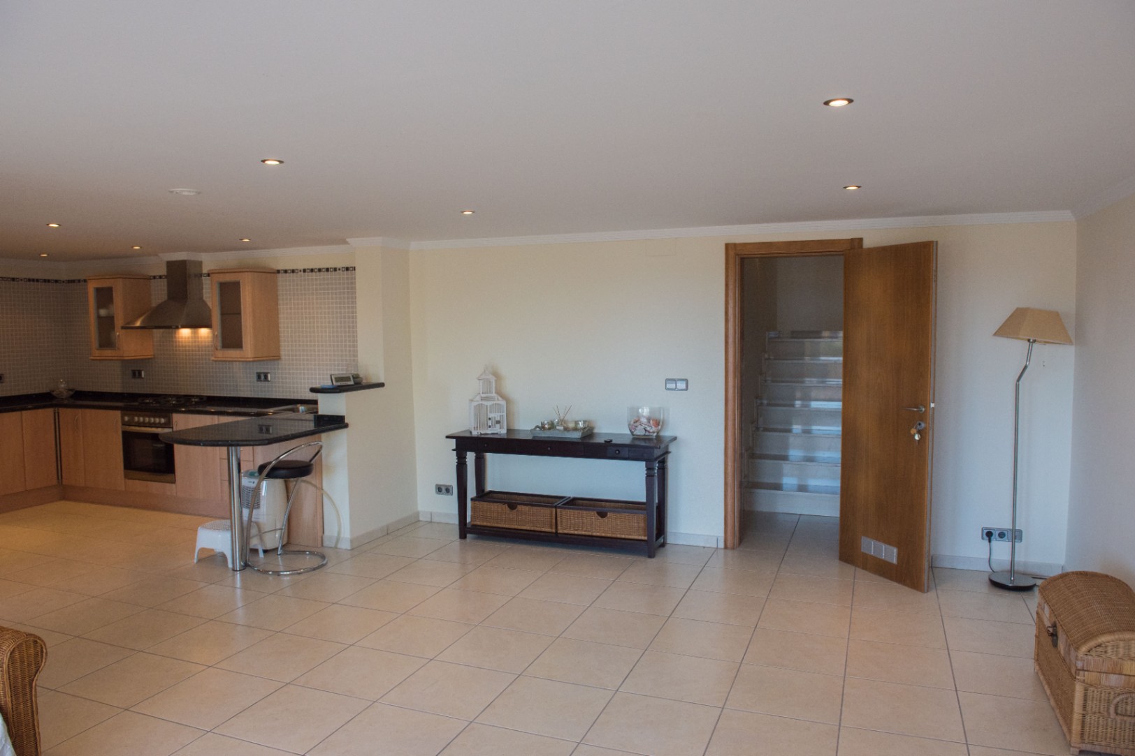 Espace de vie ouvert comprenant une cuisine avec placards en bois clair, plan de travail en granit noir et un coin repas, ouvrant sur un hall carrelé menant à l'escalier.