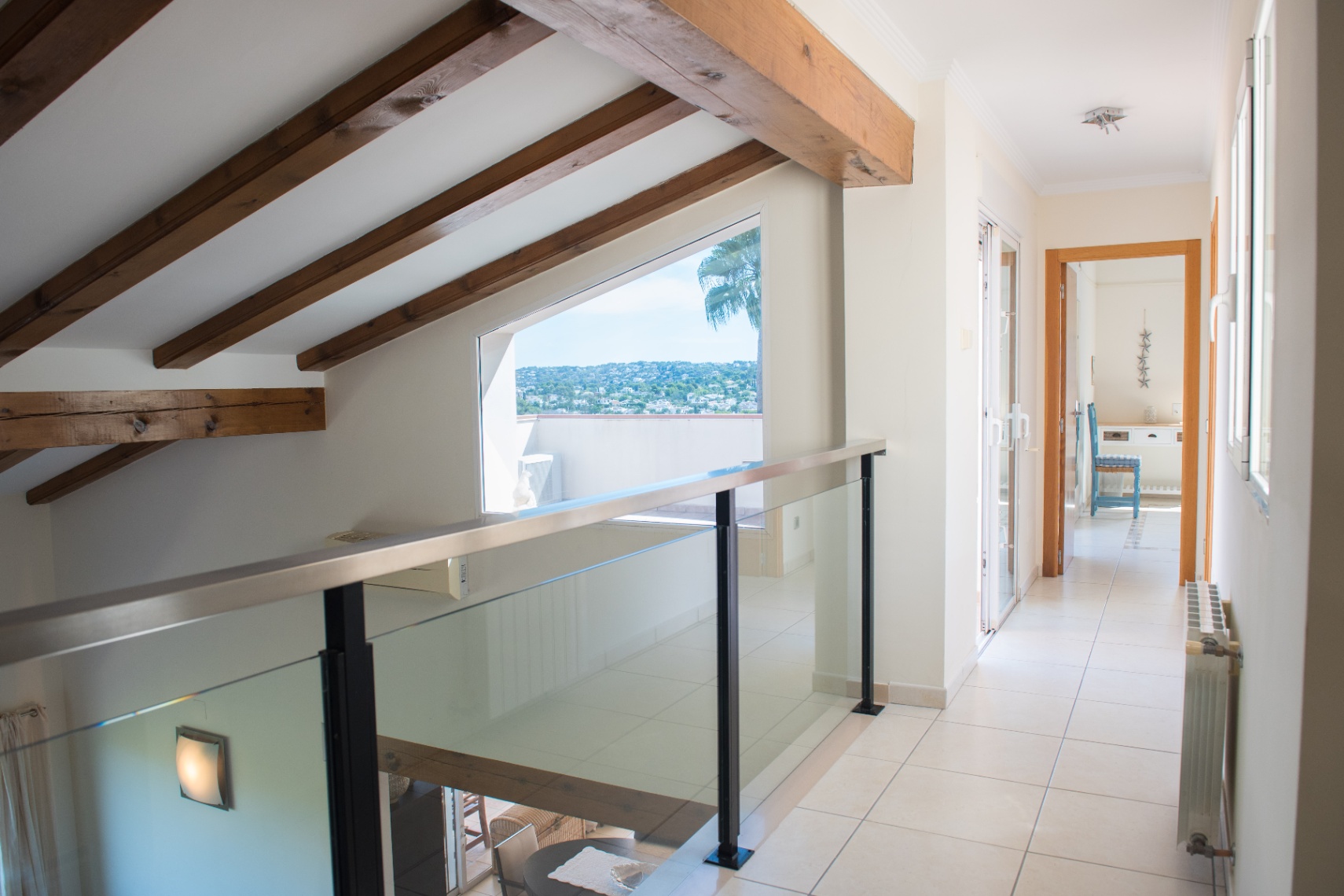Palier à l'étage avec poutres en bois apparentes, garde-corps en verre et une large fenêtre trapézoïdale offrant une vue dégagée sur le paysage.