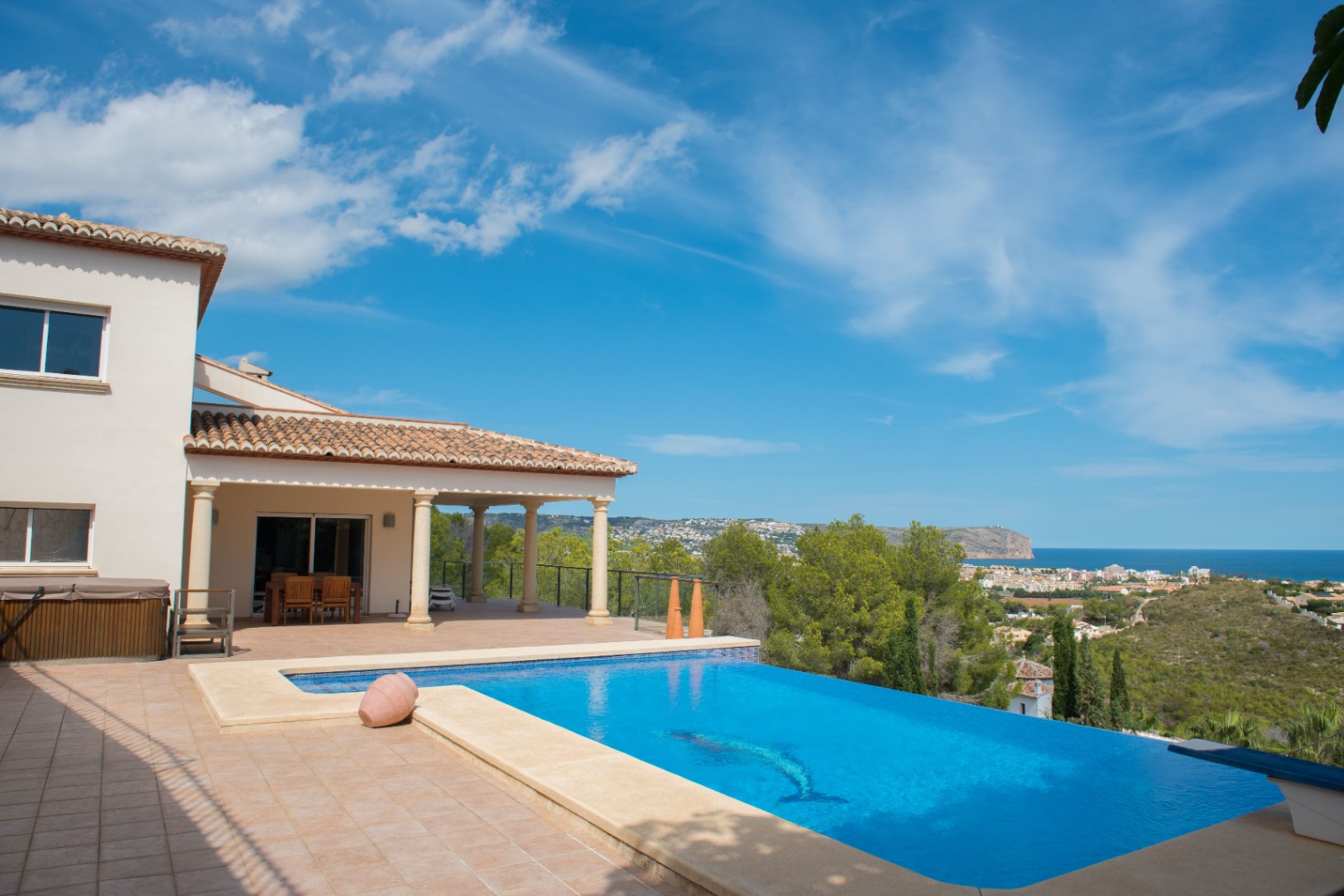 Piscine à débordement avec mosaïque de dauphin offrant une vue sur la mer. La villa comprend une terrasse couverte avec colonnes en pierre et toit en tuiles.