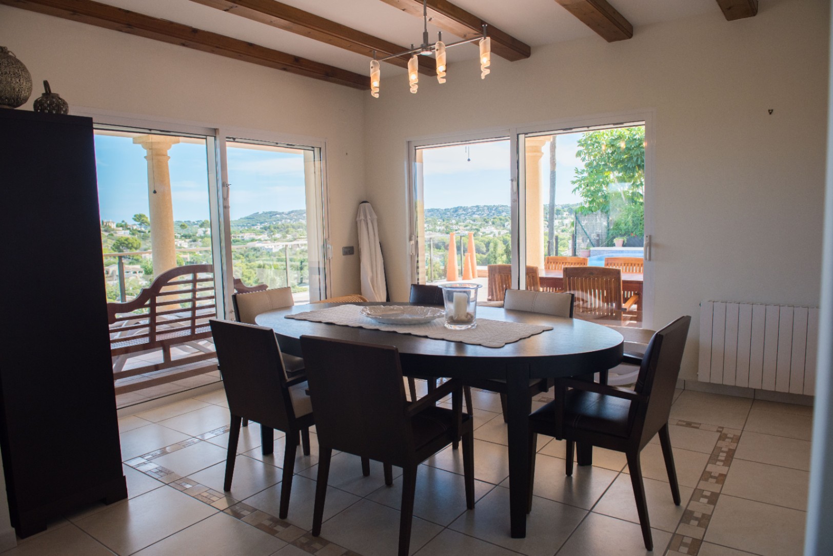 Salle à manger avec table ovale en bois sombre et poutres apparentes. De grandes baies vitrées offrent une vue panoramique sur la vallée et un accès direct à la terrasse.