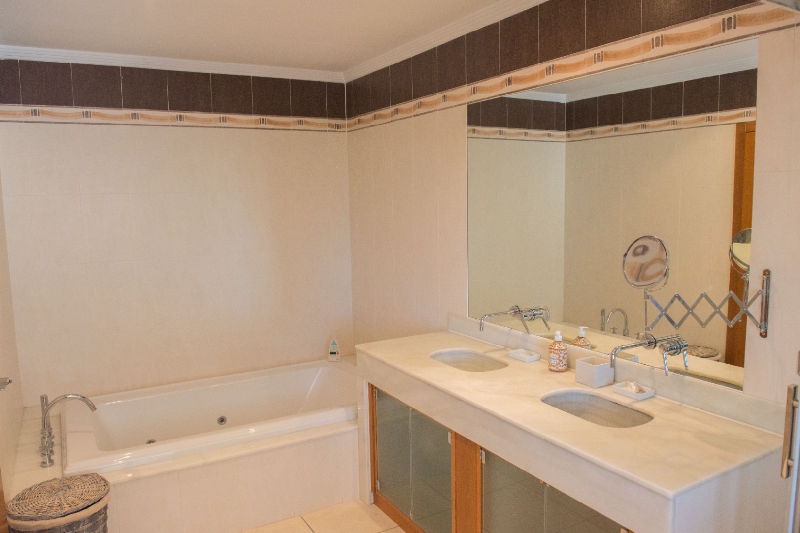 Salle de bain avec double vasque en pierre, robinetterie murale et baignoire balnéo encastrée, agrémentée d'un carrelage mural avec frise décorative.