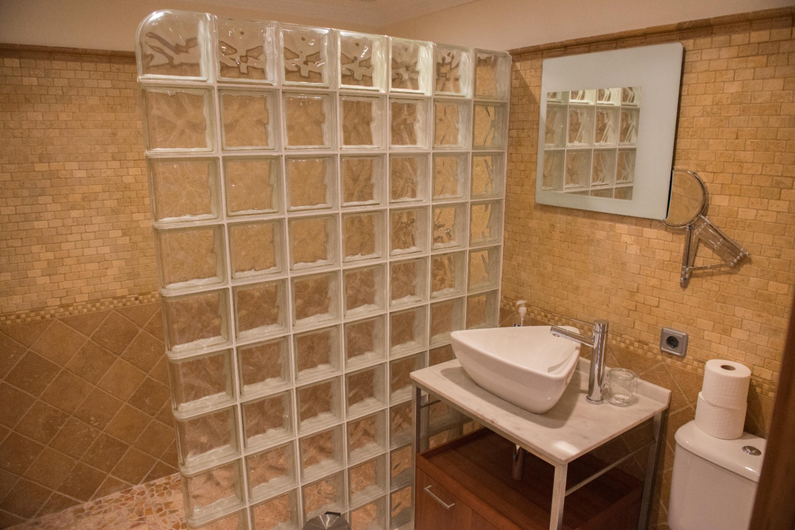 Salle de bain avec paroi de douche en briques de verre, murs en mosaïque beige et vasque à poser sur plan en marbre avec meuble en bois.