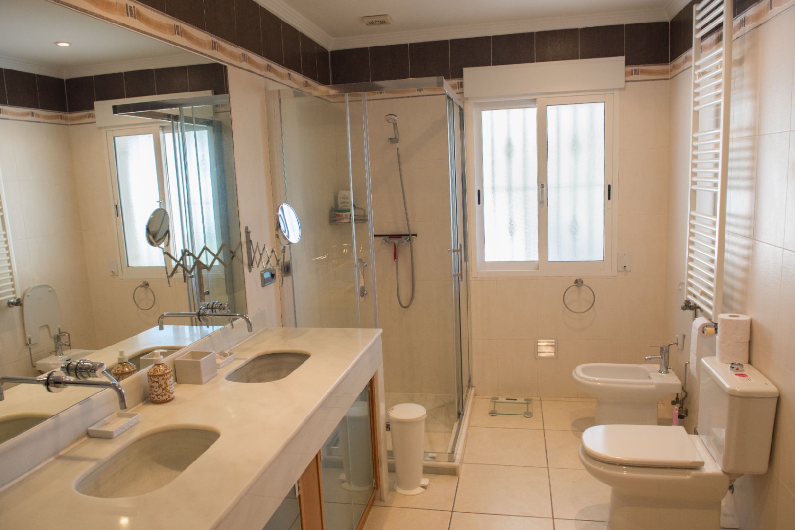 Salle de bain équipée d'un double plan vasque en marbre, d'une douche vitrée et de sanitaires blancs sur un carrelage aux tons clairs avec miroir mural.