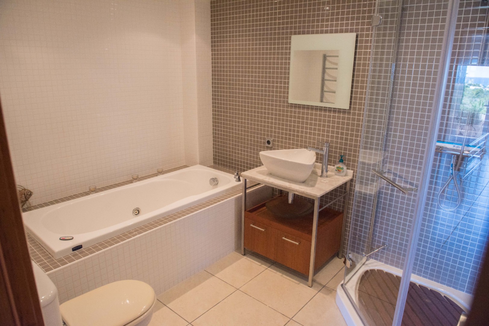Salle de bain moderne avec baignoire encastrée, douche vitrée au sol en bois et vasque à poser sur un meuble en marbre avec mosaïque taupe.
