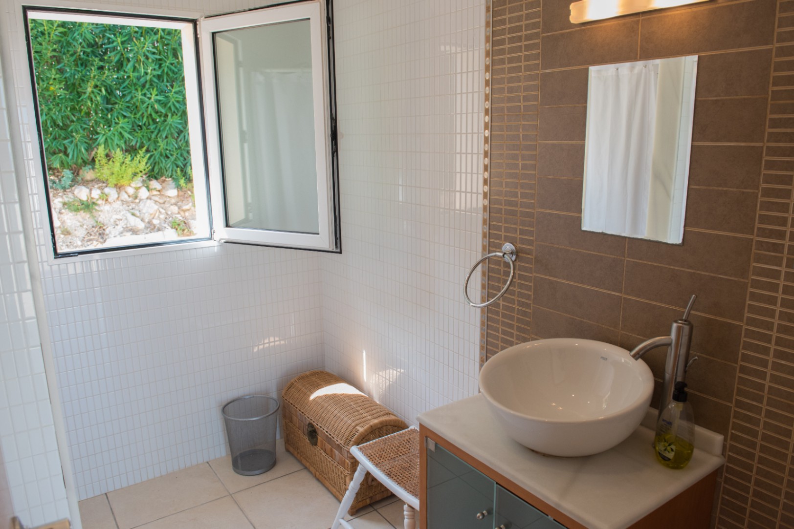 Salle de bain moderne avec vasque à poser, mur d'accent en carreaux bruns et fenêtre ouverte offrant une vue sur la végétation extérieure.