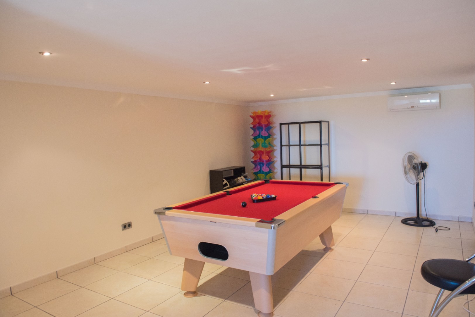 Salle de jeux spacieuse avec billard au tapis rouge, sol en carrelage clair, éclairage encastré au plafond et climatisation murale.
