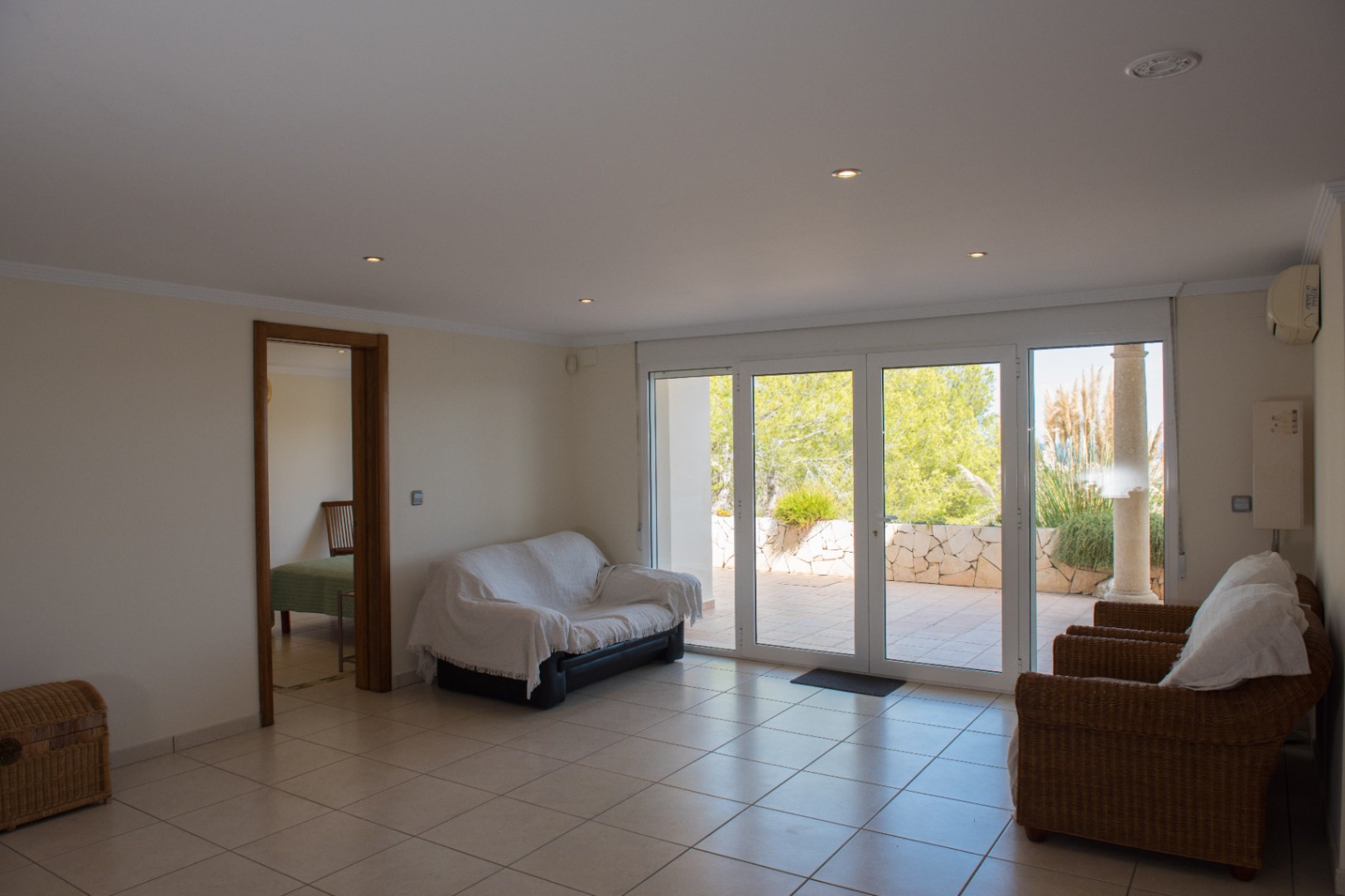 Salon avec carrelage clair et grandes baies vitrées ouvrant sur une terrasse. Comprend des fauteuils en osier, des spots encastrés et une vue sur la végétation.