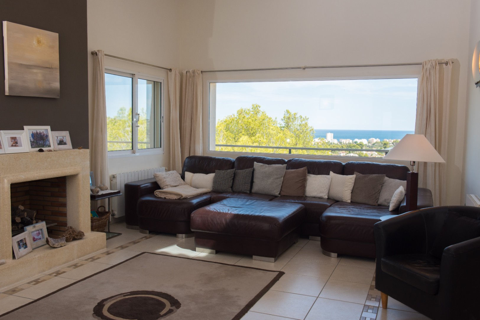 Salon spacieux avec canapé d'angle en cuir marron, cheminée en pierre et grande fenêtre panoramique offrant une vue dégagée sur la mer et le littoral.
