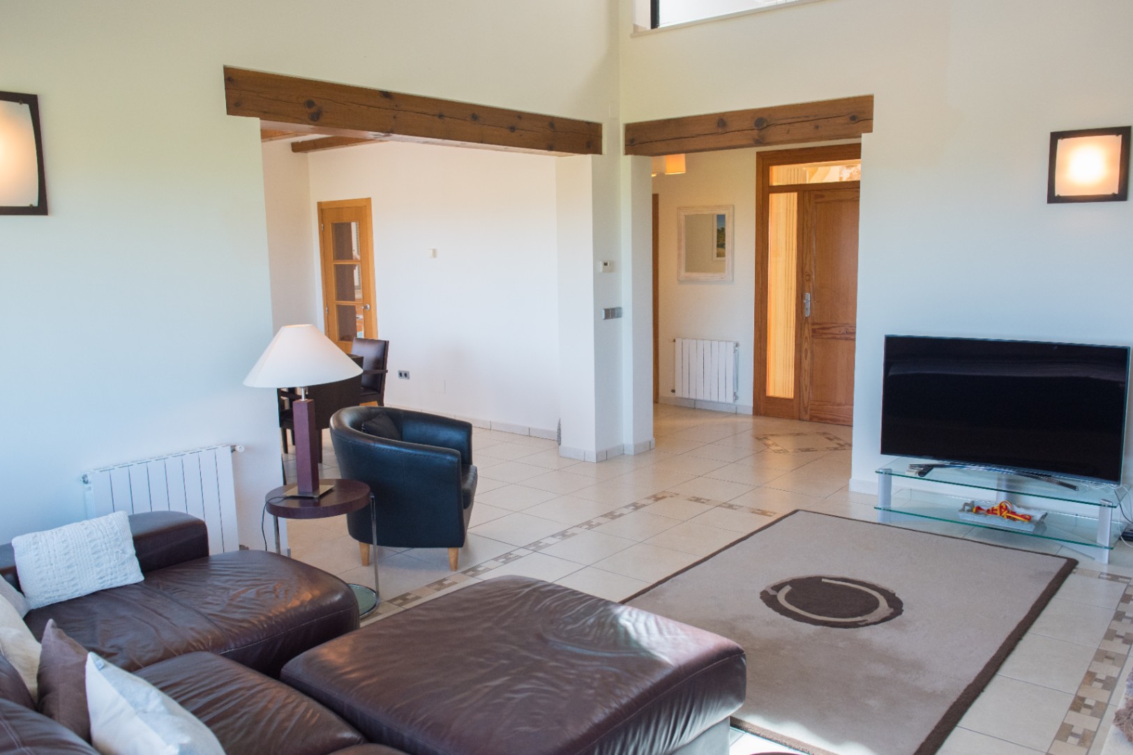Séjour avec canapés en cuir sombre, sol en céramique avec frises et poutres en bois apparentes. La disposition ouverte mène à la salle à manger et aux portes en bois.