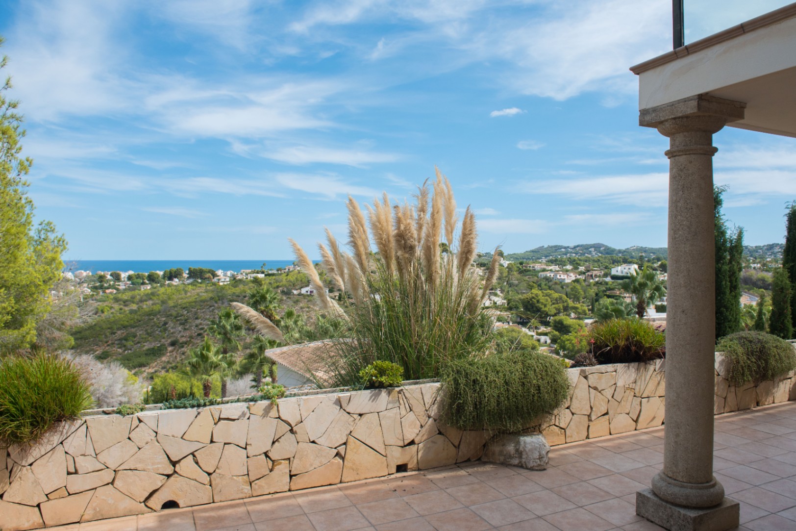 Terrasse avec colonnes en pierre et muret en pierre sèche, agrémentée d'herbes de la pampa offrant une vue panoramique sur le littoral méditerranéen.