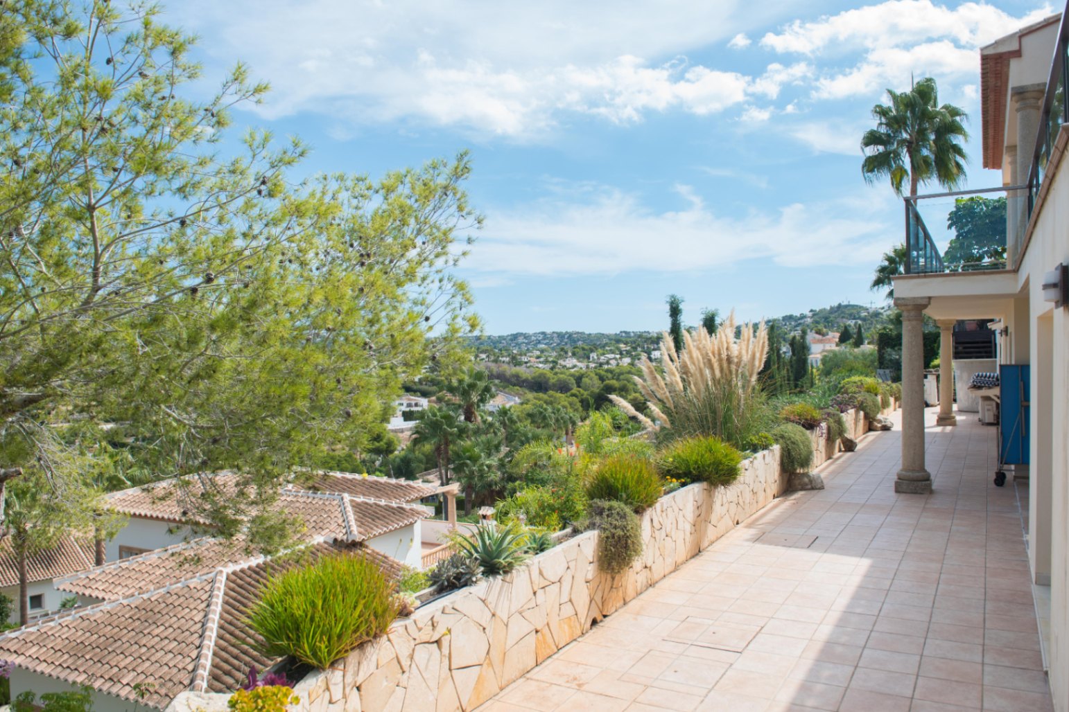 Terrasse surélevée avec carrelage beige et muret de plantation en pierre. Vue panoramique sur la vallée, les villas environnantes et la végétation locale.