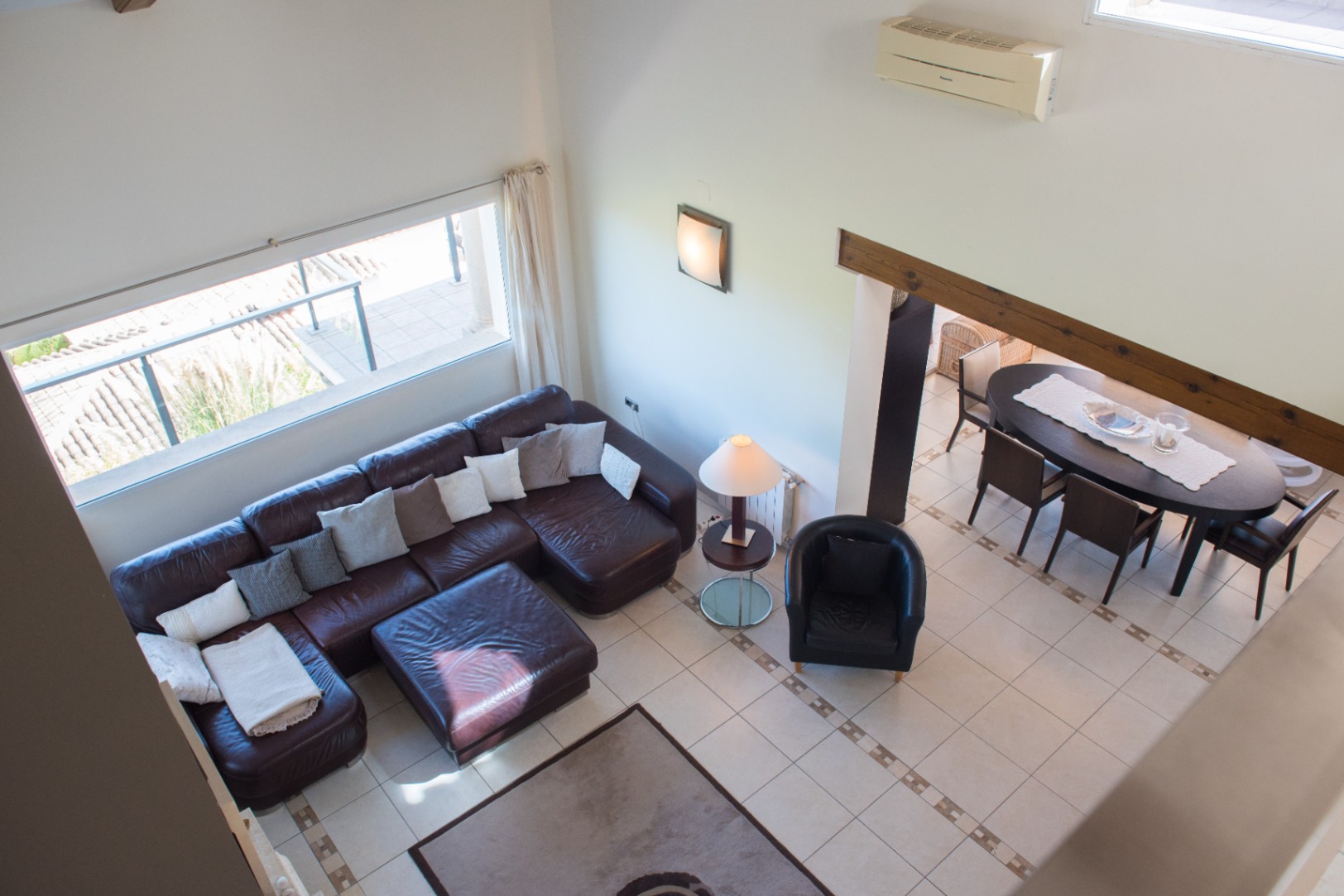 Vue en surplomb d'un salon avec canapé d'angle en cuir marron, sol carrelé et coin repas adjacent comprenant une table ovale sous une poutre en bois apparente.