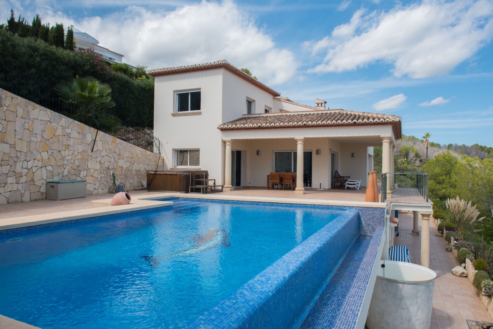 Vue extérieure d'une villa blanche avec piscine à débordement en mosaïque bleue et terrasse couverte soutenue par des colonnes classiques.