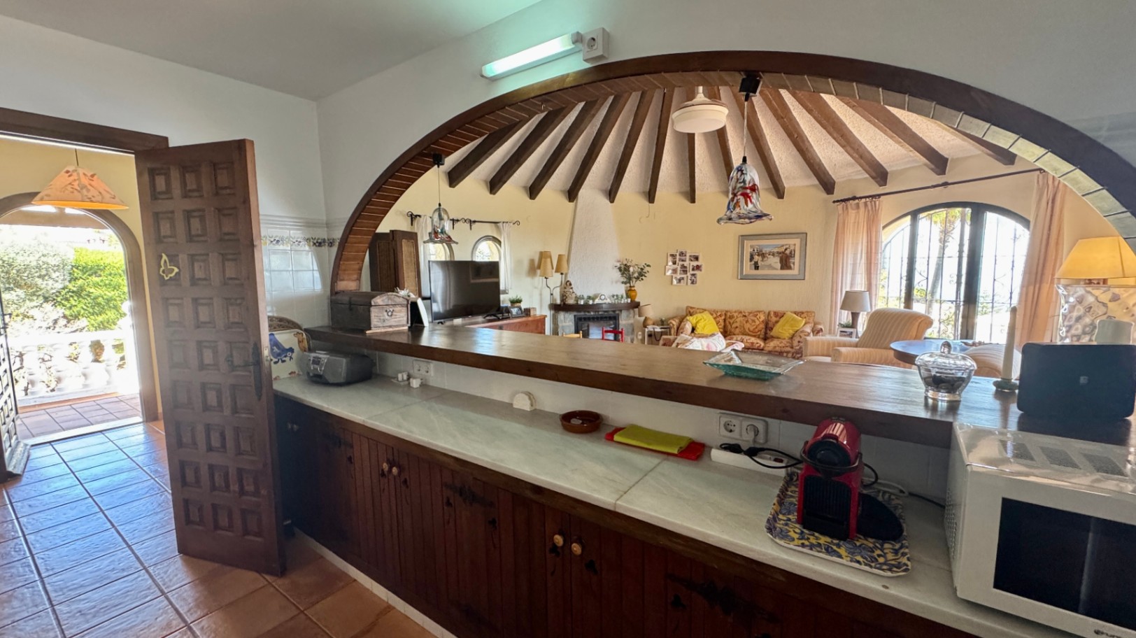 Bar de cuisine avec plan de travail en marbre, s'ouvrant par une arche en bois sur un salon circulaire doté d'un plafond à poutres radiales.