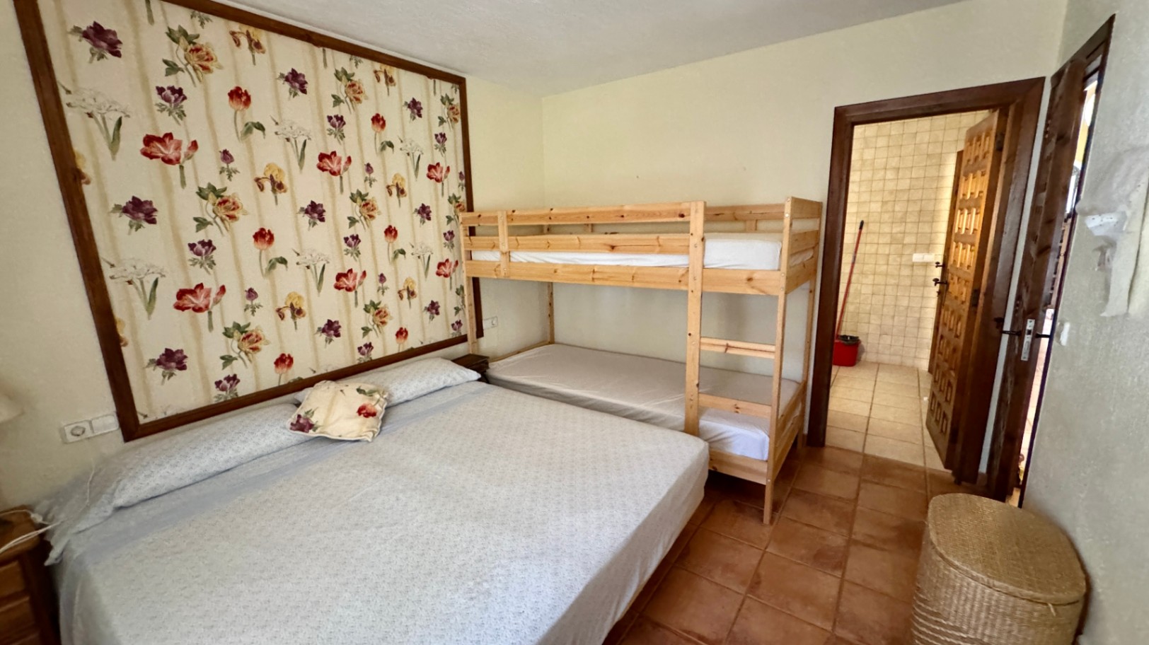 Chambre avec lit double et lits superposés en bois, agrémentée d'un panneau mural floral et d'un sol en terre cuite. Accès direct à une salle de bain.