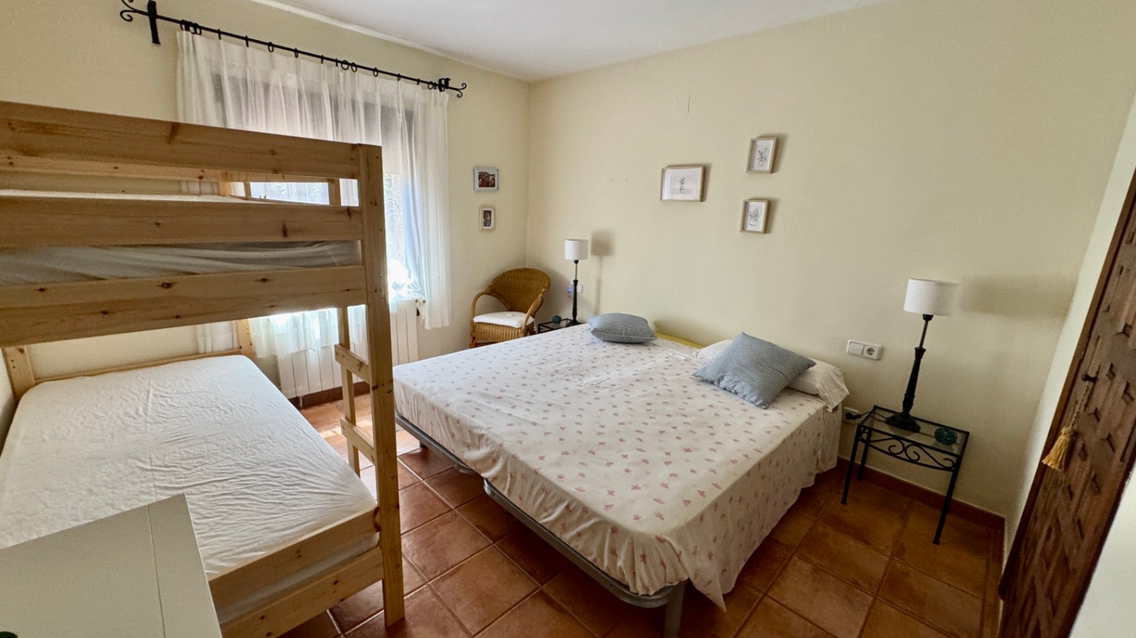 Chambre avec lits superposés en bois et lit double, sol en terre cuite et lumière naturelle. Équipée d'un fauteuil en osier et d'une décoration murale sobre.