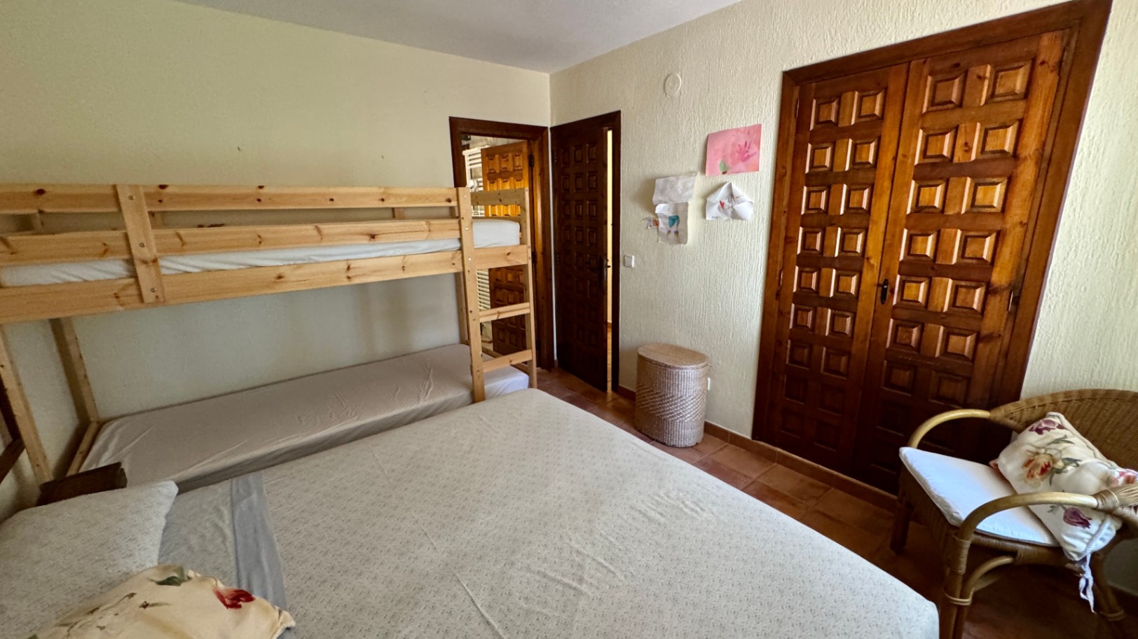 Chambre avec lits superposés en pin, armoire encastrée en bois à panneaux et sol en terre cuite. Comprend un fauteuil en osier et des murs à crépi.