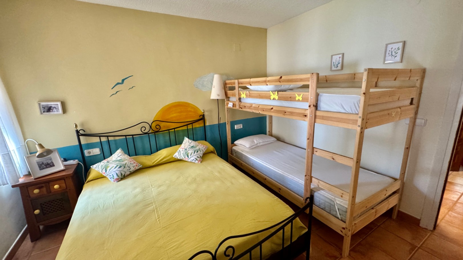 Chambre équipée d'un lit double en fer forgé et de lits superposés en bois naturel. Elle dispose d'un sol en terre cuite et d'une fresque murale thématique.