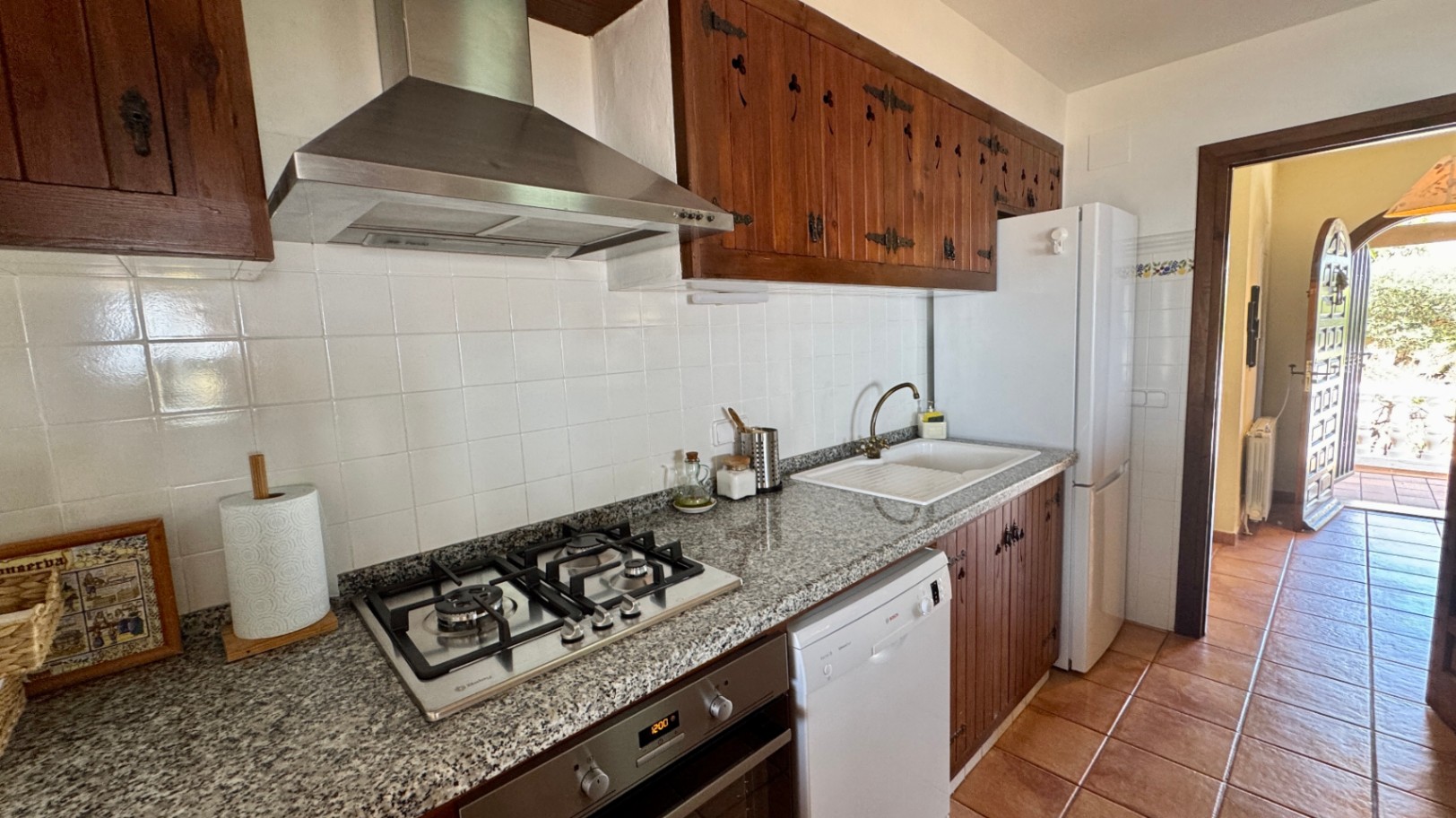 Cuisine rustique avec meubles en bois sombre, plan de travail en granit et cuisinière à gaz. Sol en terre cuite et crédence en carrelage blanc avec électroménagers blancs.