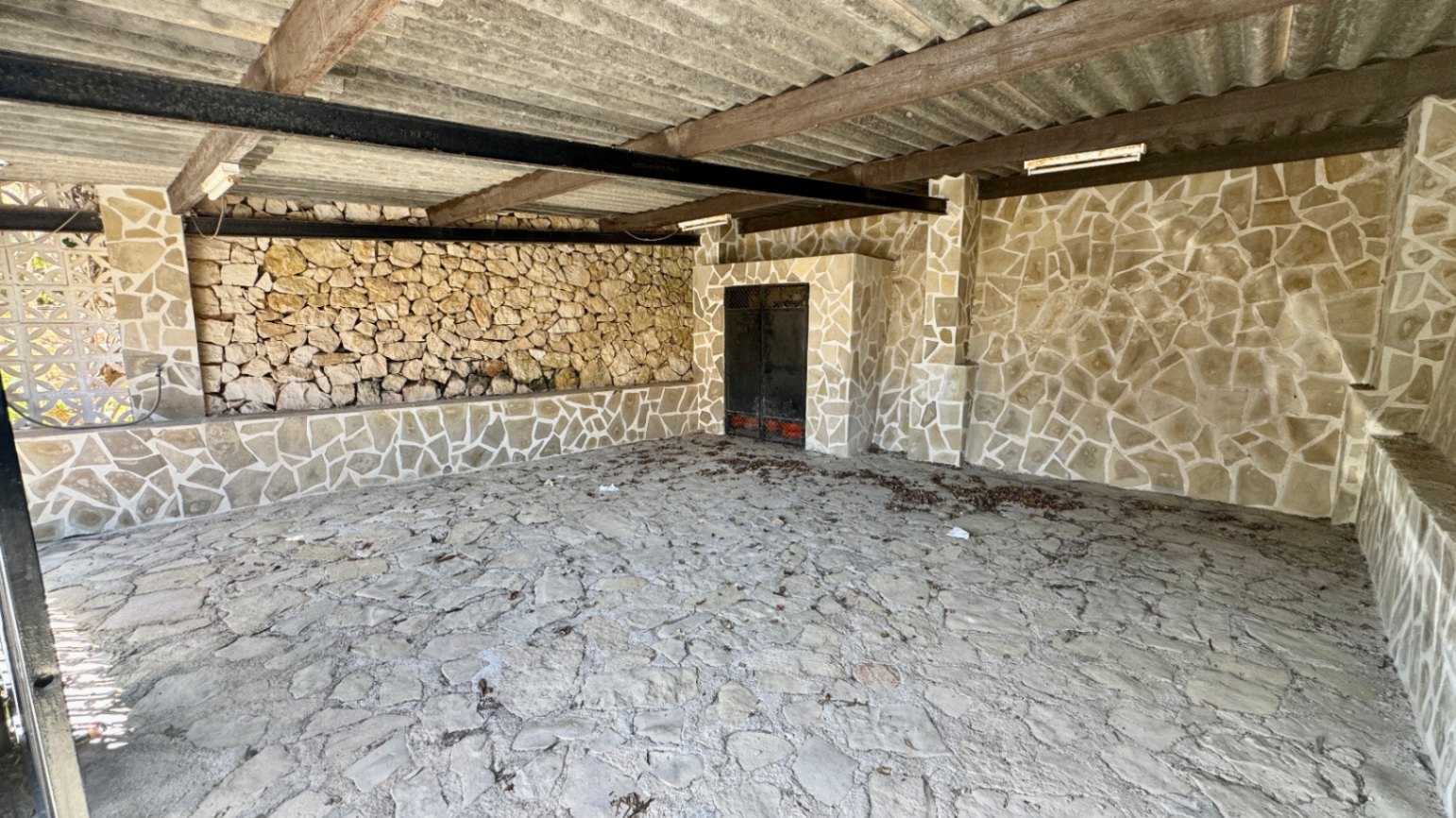 Espace extérieur couvert avec murs et sol en pierre apparente, structure à poutres en bois et métal sous un toit en tôle ondulée.